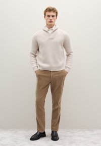 Hellbeiger, gerippter Strickpullover mit Schalkragen, kombiniert mit braunen Cordhosen und schwarzen Lederschuhen. Minimalistisches Design, figurbetonte Silhouette.