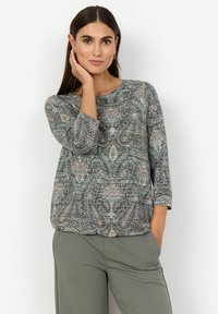Femme aux cheveux foncés portant un haut à motifs paisley avec des manches trois-quarts et un pantalon vert, posant avec une main sur son visage.