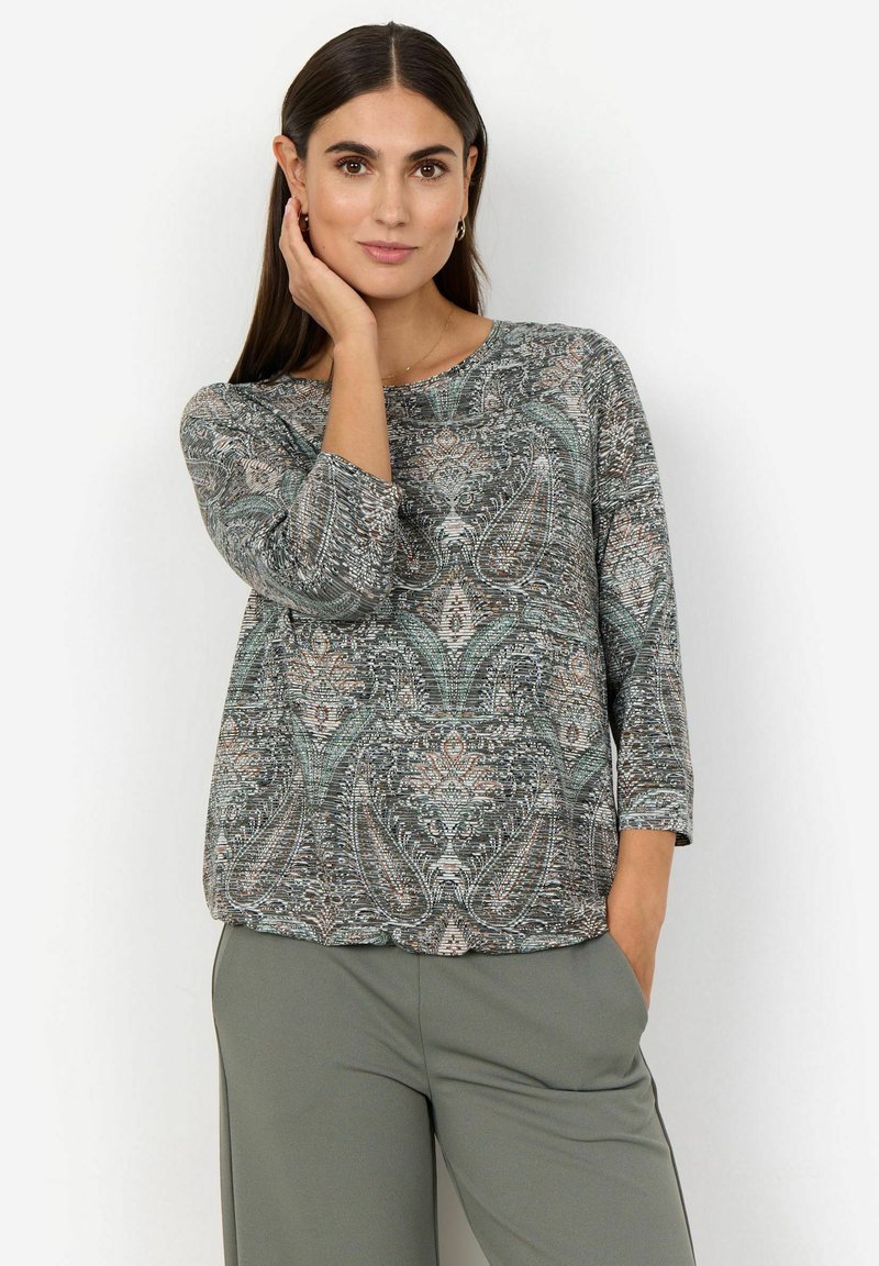 Femme aux cheveux foncés portant un haut à motifs paisley avec des manches trois-quarts et un pantalon vert, posant avec une main sur son visage.