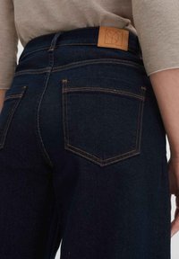 Jeans in denim scuro con vita alta, caratterizzati da cuciture arancioni e un'etichetta in pelle beige sul retro. Sono visibili due tasche posteriori.