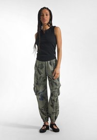 Top negro sin mangas con escote en forma de ojo de cerradura, combinado con pantalones cargo verdes con estampado floral y puños elásticos. Zapatos negros.