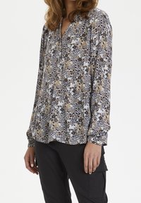 Blouse fleurie avec des motifs bleus, noirs et dorés ; manches longues, encolure en encoche, et coupe décontractée en tissu léger.