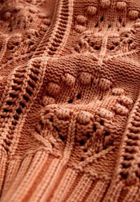 Gros plan sur un tissu tricoté doux de couleur rouille avec des motifs en losanges et points en relief ainsi que des bords côtelés.