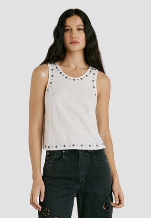 Jeune femme aux longs cheveux foncés portant un haut blanc sans manches orné de clous métalliques et un jean noir avec des découpes florales.