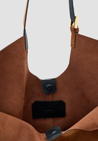 Borsa tote in camoscio marrone con interno in pelle nera, dotata di chiusura con snap magnetico e di un'etichetta in pelle nera all'interno. Spallacci regolabili.