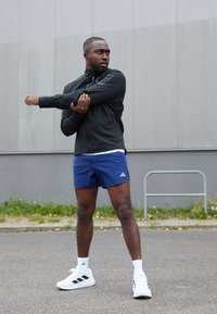 Černá sportovní bunda, modré šortky a bílé tenisky. Bunda má vysoký límec a elastický materiál; šortky jsou hladké s logem Adidas.