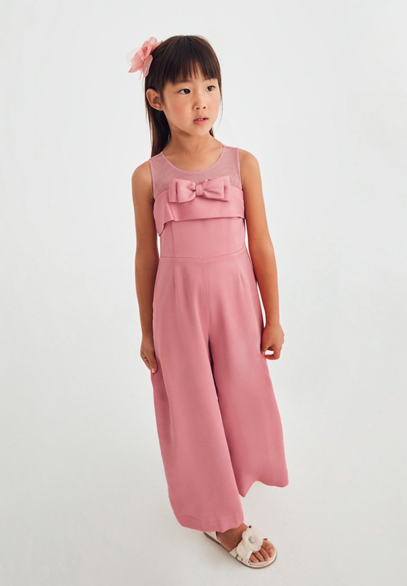 Tuta rosa con un corpo a strati e un grande fiocco. Realizzata in tessuto leggero, ha gambe ampie e una texture liscia. Sandali bianchi inclusi.