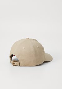Beige bomullsbasket caps med buet skygge, justerbar stropp med metallspenne, og ventilasjonslinser bak.