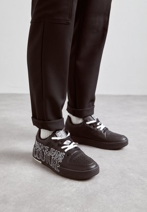 Czarne sneakersy z białymi sznurowadłami i logo „Versace Jeans Couture”, noszone z czarnymi podwiniętymi spodniami i białymi skarpetkami na szarej podłodze.
