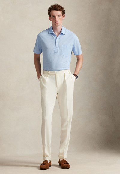 Ανοιχτό μπλε πόλο μπλουζάκι με τσέπη, συνδυασμένο με κρεμ παντελόνι και καφέ σουέντ μοκασίνια. Η εμφάνιση διαθέτει κλασική, εφαρμοστή γραμμή.