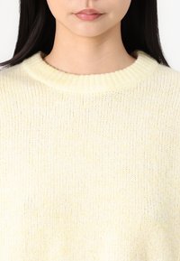 Hellgelber gestrickter Pullover mit geripptem Kragen, der eine weiche Textur und eine lässige Passform im Brustbereich aufweist.