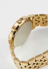 Gold metal watch with a round case, textured bezel, crown, and a linked bracelet band. The case back is smooth and reflective.