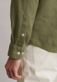 Green linen shirt sleeve featuring cuffs with cream buttons and a smooth texture, paired with light-colored pants.