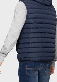 Marineblaue gepolsterte Weste mit horizontalen Nähten, Kapuze und Fronttaschen, getragen über einem grauen Langarmshirt und Jeans.