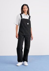 Mustad denim kombinesed, millel on reguleeritavad sangad, eesmine tasku ja logoga plaaster, koos valge triibulise särgi ja valgete tossudega.