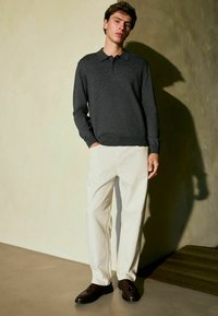 Dunkelgraues, langärmliges Poloshirt mit Kragen, kombiniert mit locker sitzenden, cremefarbenen Hosen und dunkelbraunen Schuhen, stehend vor einer strukturierten Wand.