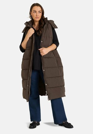 Femme debout portant un long manteau sans manches matelassé marron sur un haut noir, un jean bleu et des chaussures noires sur un fond uni.
