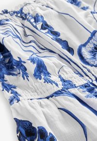 White fabric featuring blue floral patterns, with gathered detailing along the top edge and flowing texture. Lightweight and soft appearance.