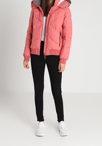 Veste matelassée rose avec capuche détachable et bordure en fourrure. Fermeture éclair à l'avant, poches latérales, portée avec un pantalon noir et des baskets blanches.