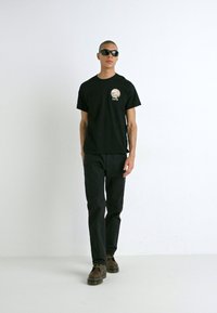 Un tricou negru cu un logo rotund Levi's, purtat cu pantaloni negri. Modelul poartă ochelari de soare negri și pantofi maro, pe un fundal alb simplu.