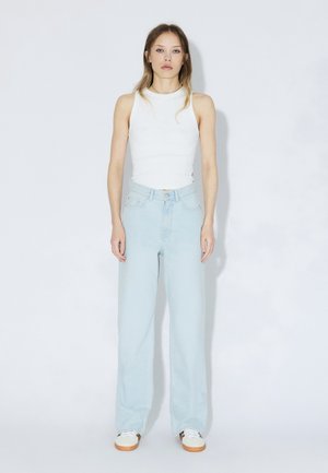 Jeans azzurri a vita alta con vestibilità ampia, abbinati a una canotta bianca aderente. Sneakers bianche con suole marrone chiaro.