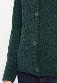 Cardigan en tricot vert foncé avec un motif en losanges, des poignets côtelés et cinq boutons assortis sur le devant. Détail de tissu texturé visible.