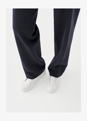 Valkoiset tennarit, joissa on kuvioidut kumipohjat ja litteät nauhat, yhdistettynä tummansinisiin housuihin, joiden kangas on löysä ja sileä sekä istuvuus räätälöity.