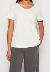 Camiseta blanca de cuello en V de tejido suave con mangas cortas. Presenta filas verticales de adornos plateados a lo largo del lado izquierdo.