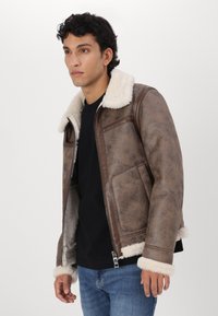 Ruda dirbtinė odinė striukė su avių kailio apykakle ir rankogaliais, turinti užtrauktuką ir priekinius kišenes. Textūruotas paviršius su senoviniu įvaizdžiu.