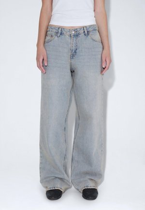 Jean large en denim bleu clair, doté d'un design classique à cinq poches et d'une finition légèrement délavée. Porté avec des chaussures noires.