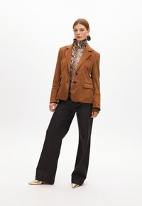 Hnědý semišový blazer se dvěma knoflíky, bočními kapsami, nošený přes černý a béžový vzorovaný top. Doplňují ho široké tmavé kalhoty a zlaté boty.