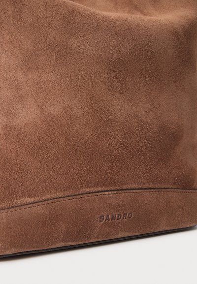 Gros plan d'un tissu en daim marron avec des coutures visibles et la marque "SANDRO" embossée près du bord inférieur.