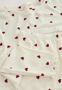 Two-piece loungewear set in cream with ribbed texture, decorated with small maroon hearts. Features a round neck and ruffled sleeve cuffs.
