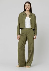Veste courte vert olive en lin, avec deux poches avant et fermeture à boutons ; associée à un pantalon large et un top blanc texturé.