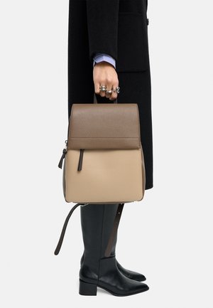 Osoba w czarnym płaszczu i butach trzyma beżowo-brązowy skórzany plecak za górną rączkę, pokazując wiele srebrnych pierścieni na dłoni.