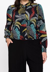 Blusa de botones en tela oscura con patrones florales coloridos. Cuenta con un collar, mangas largas y un corte relajado. Combinada con pantalones negros.