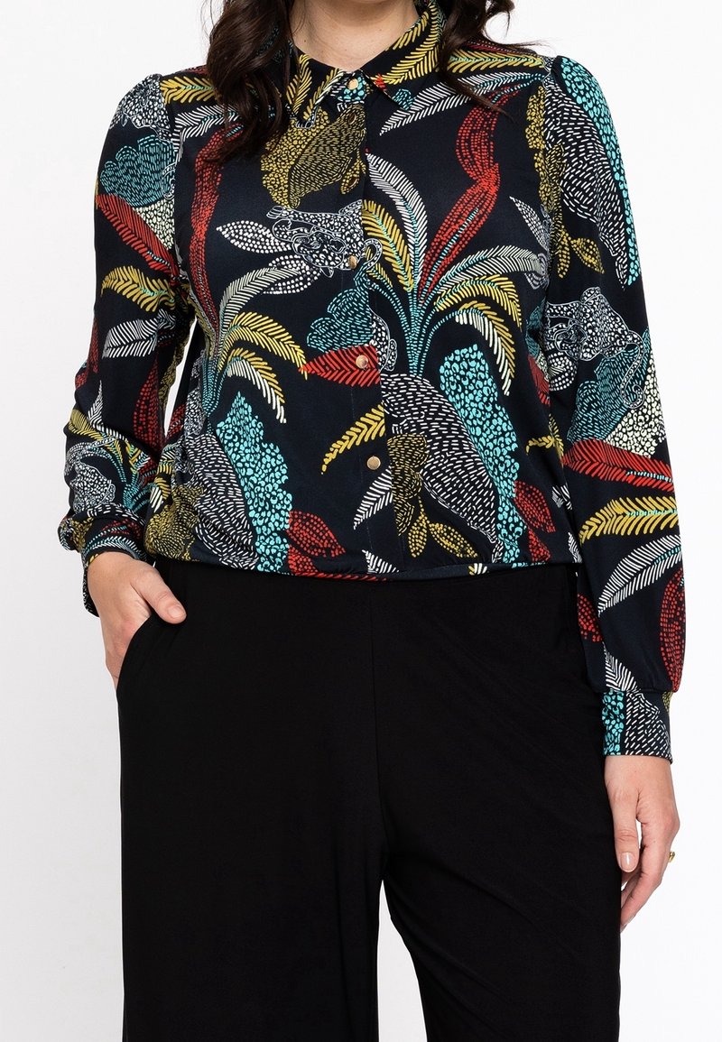 Blusa de botones en tela oscura con patrones florales coloridos. Cuenta con un collar, mangas largas y un corte relajado. Combinada con pantalones negros.