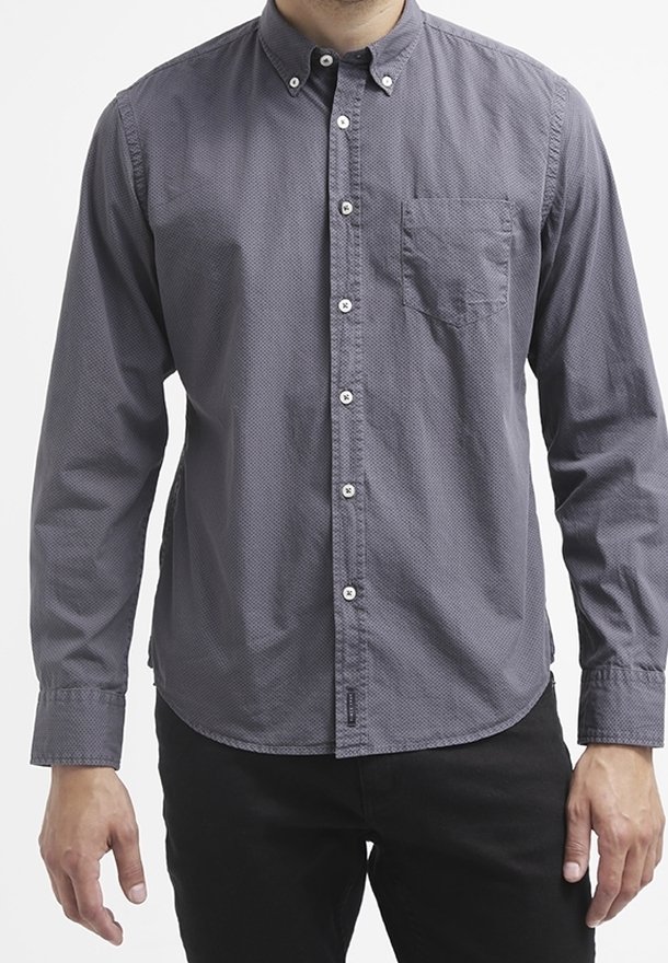 Homme portant une chemise à manches longues gris foncé avec une poche poitrine et un pantalon noir, debout les bras détendus.