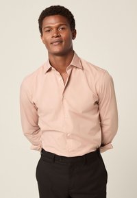 Hellrosa Hemd mit Knopfleiste, spitzem Kragen, figurbetontem Schnitt und langen Ärmeln. Kombiniert mit schwarzer Hose ergibt sich ein insgesamt eleganter Look.