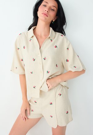 Femme aux longs cheveux foncés portant une chemise beige à manches courtes boutonnée et un short assorti avec de petits motifs floraux rouges.