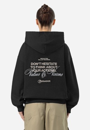 Osoba z warkoczami związanymi w kok, ubrana w czarną bluzę z napisem "Nie wahaj się myśleć o swoich czynach" i beżowe spodnie, stojąca tyłem.