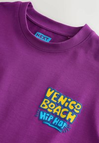 Lila, kerek nyakú póló "Venice Beach Hip Hop" logóval, vastag sárga és kék betűkkel a mellrészen, valamint kék "NEXT" márkajelzés a gallér belsejében.