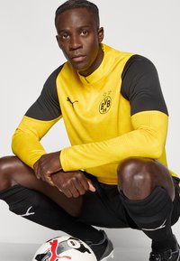 Αθλητική φανέλα με μακριά μανίκια και φερμουάρ, σε κίτρινο και μαύρο χρώμα, με το λογότυπο BVB 09 και υφή σχεδίου. Μαύρο σορτς και κάλτσες.