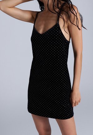 Vrouw in een mouwloze zwarte minijurk met zilverkleurige stippen, poserend met één arm omhoog en nat haar tegen een effen achtergrond.