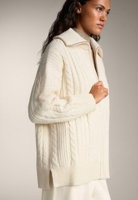 Cardigan en grosses mailles avec un col, fermeture éclair et poignets côtelés. Présente des motifs texturés et une coupe décontractée.