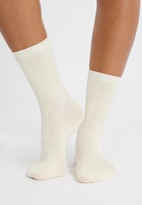 Un paio di calzini leggeri color beige indossati a piedi nudi, un piede piatto e l'altro leggermente sollevato che mostra il tallone.