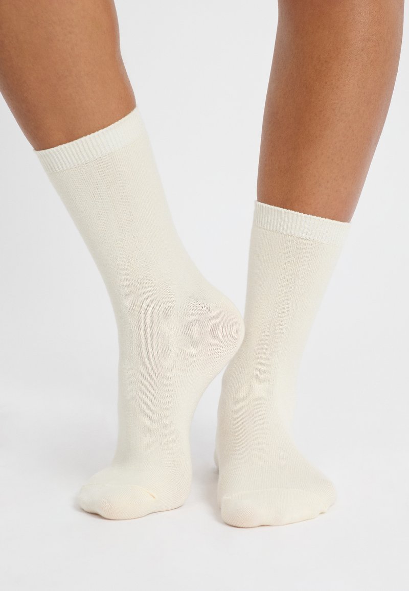 Un paio di calzini leggeri color beige indossati a piedi nudi, un piede piatto e l'altro leggermente sollevato che mostra il tallone.