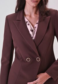 Brauner zweireihiger Blazer in strukturiertem Design mit goldenen, runden Akzenten, kombiniert mit einer hellrosa Bluse mit dunklen Pünktchen.