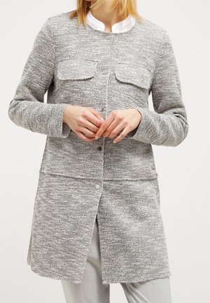 Femme portant une longue veste en tricot gris texturé avec des boutons-pression et des poches à rabat, par-dessus une chemise blanche et un pantalon gris clair.