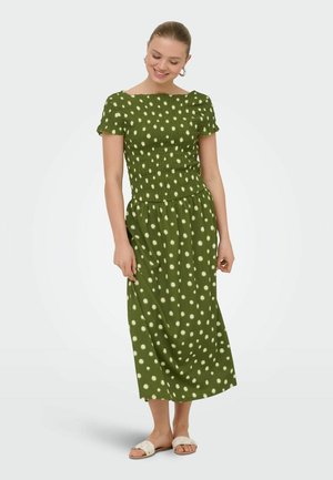 Kobieta uśmiechnięta, ubrana w zieloną midi sukienkę z krótkim rękawem w białe kropki oraz białe płaskie sandały, stoi na jednolitym tle.